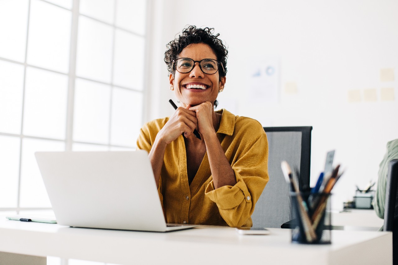 Happy graphics designer sitting at her desk thinking about a design. Business woman smiling as she contemplates an idea. Creative woman working with a laptop and a stylus pen in an office.