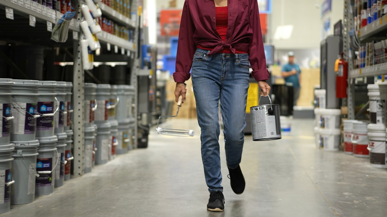 Woman shown from waist down, carrying can of paint and roller walking down paint aisle in hardware store
